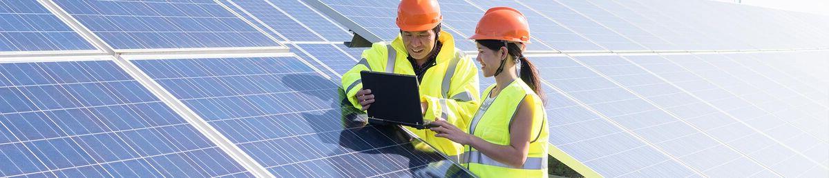 Two people in yellow hi-vis vests 和 orange safety helmets leaning on a solar panel in a field, 看iPad
